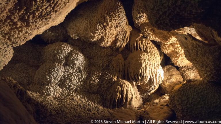 Hanging and rounded cave formations overlap under amber light in a dark cavern.