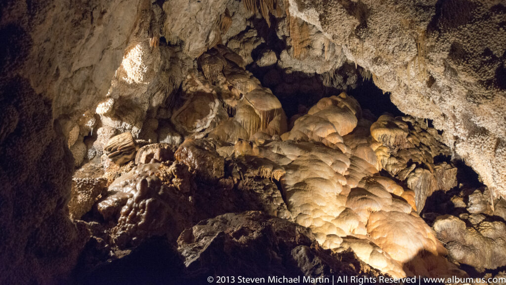 A broad cave chamber contains pale rounded deposits and sloping flowstone under spotlights.