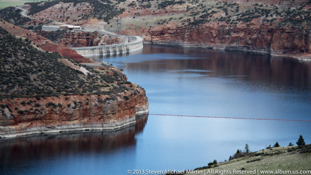 A concrete dam spans a blue reservoir between layered red-rock canyon walls.
