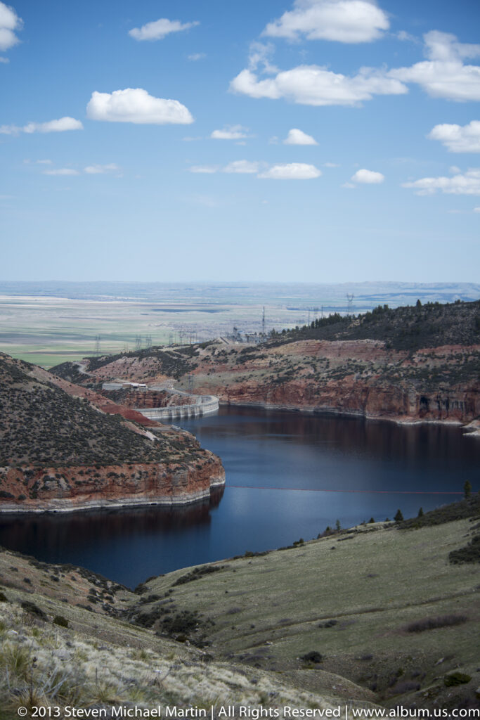 A reservoir curves through red-rock hills toward a broad plain and distant fields.