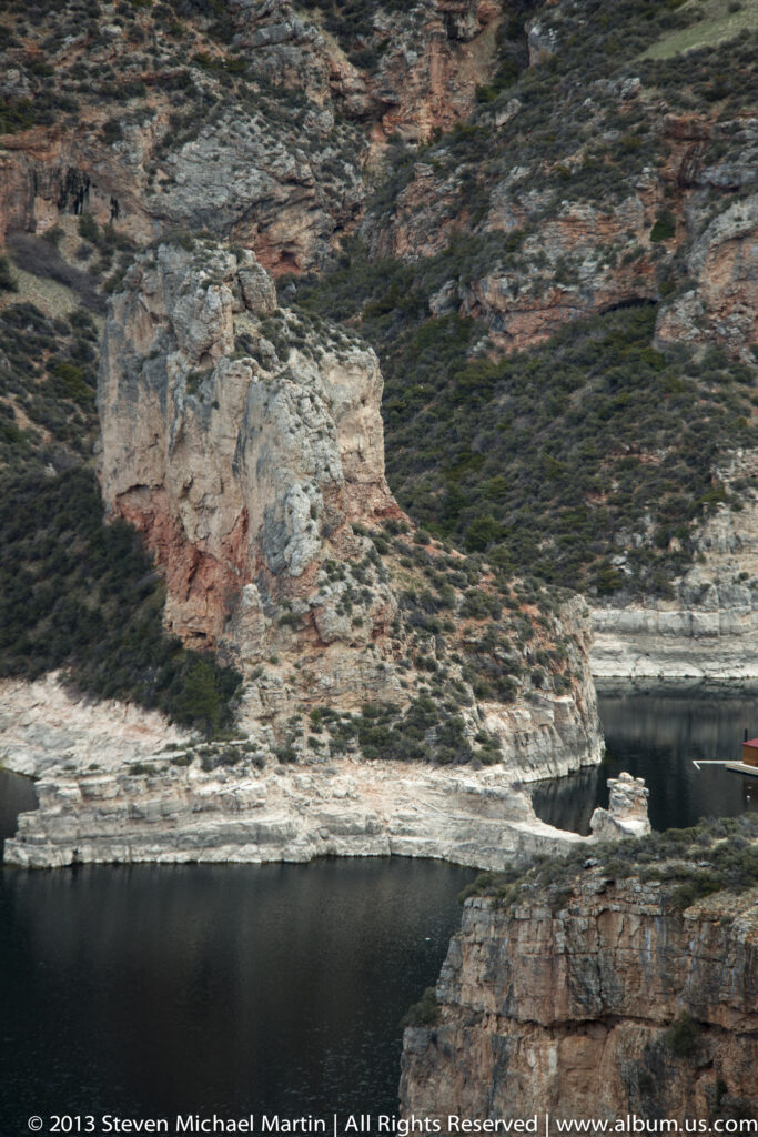 Layered pale cliffs and red rock rise above dark reservoir water along a shrub-covered canyon slope.