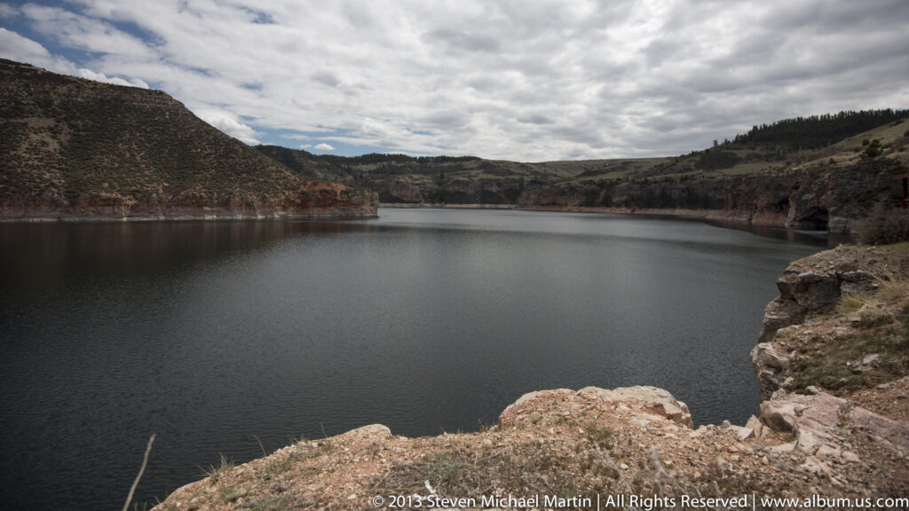 Still reservoir water sits beneath an overcast sky, bordered by rocky slopes and a rough shore.