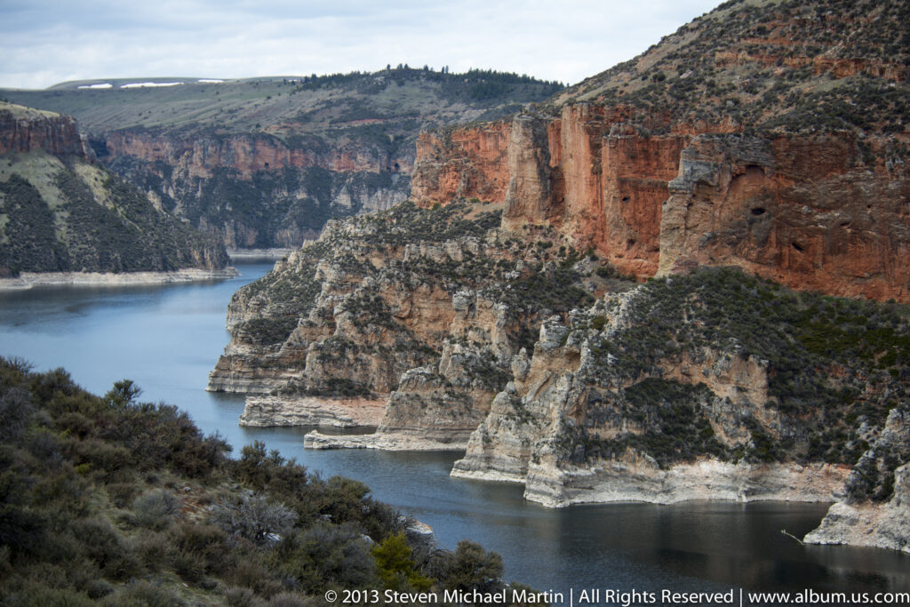 Blue water winds between red upper cliffs, pale rock ledges, and a shrub-covered foreground slope.