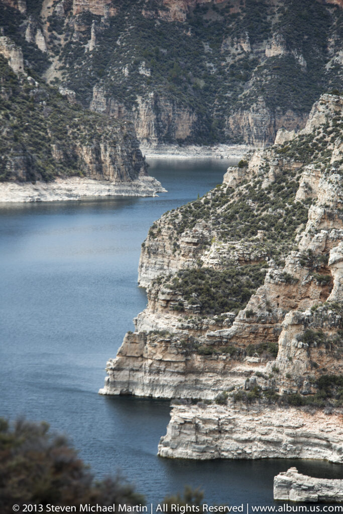 A pale stratified rock point projects into blue water beneath dark, shrub-covered canyon slopes.