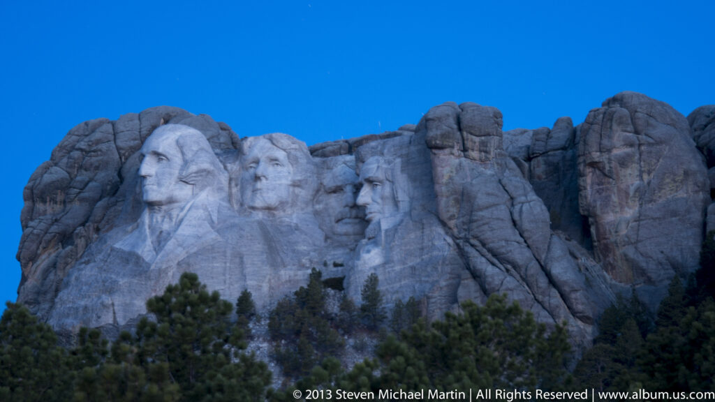 The four presidential heads at Mount Rushmore rise above dark evergreen trees in cool evening light.