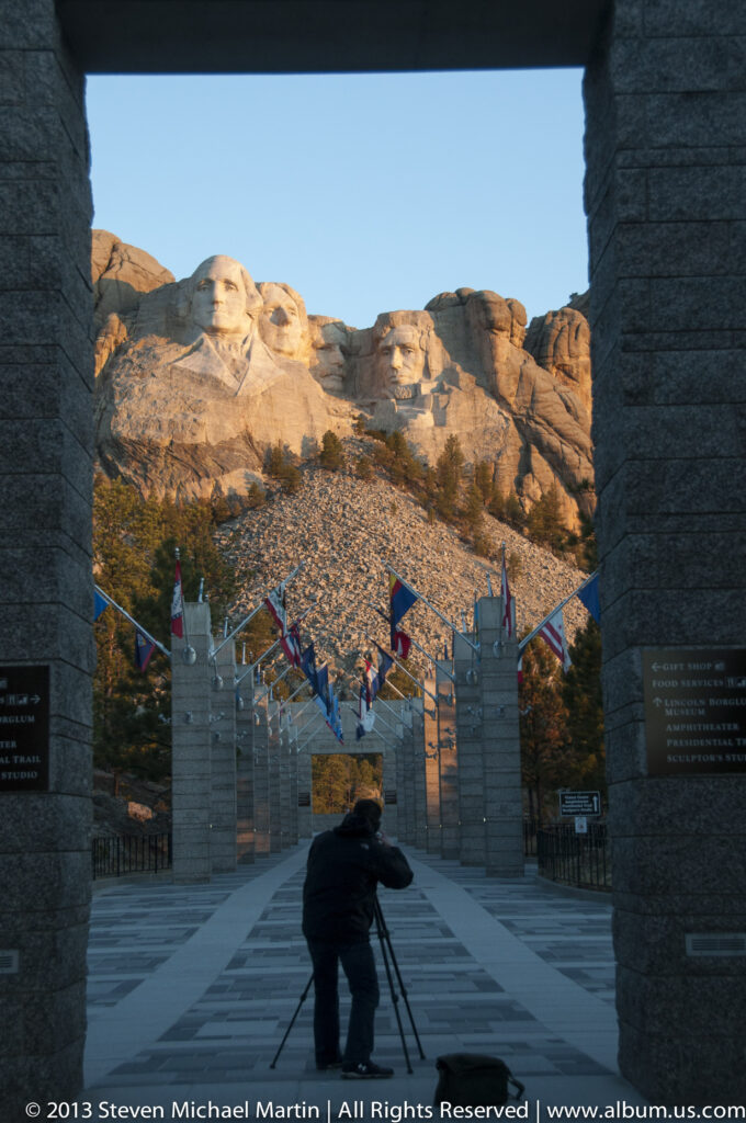 A photographer with a tripod faces Mount Rushmore from a shadowed stone passage.