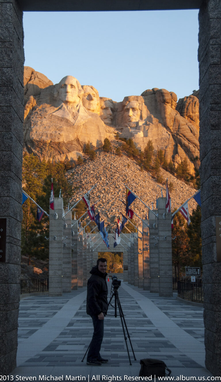 A visitor carrying camera equipment crosses the flag-lined plaza beneath Mount Rushmore.