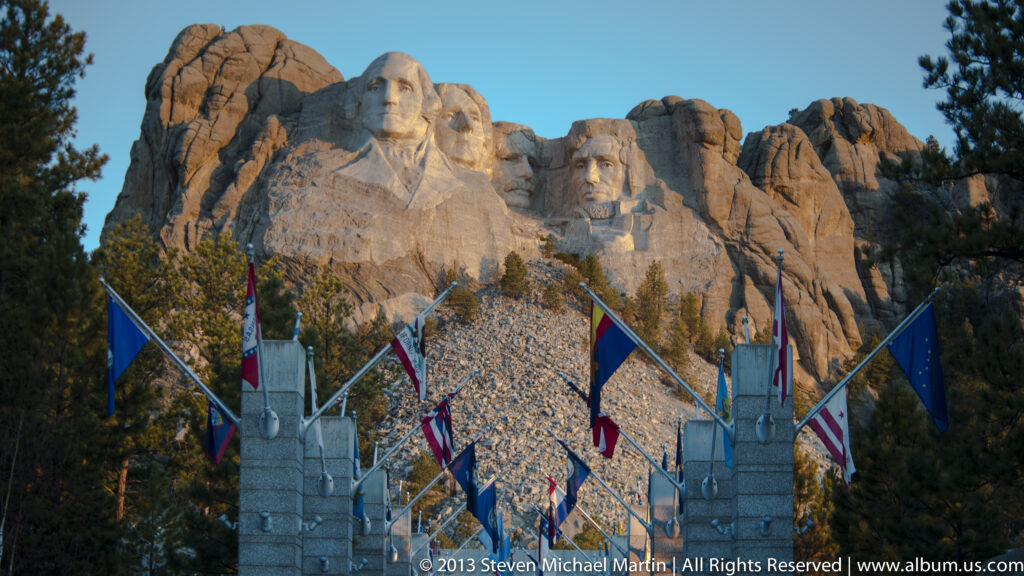 A central flag-lined path leads through stone posts toward Mount Rushmore at dusk.