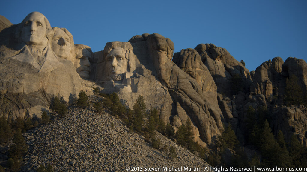 The four carved presidents span a granite cliff above a steep slope of broken rock and scattered trees.