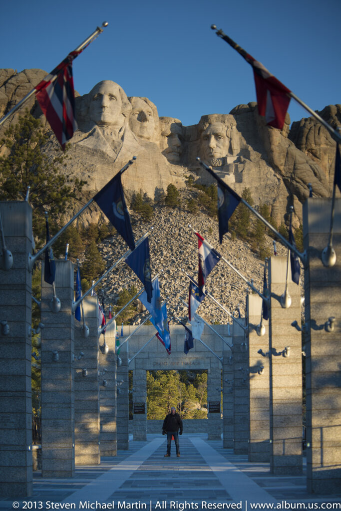 Mount Rushmore rises above a flag-lined stone entrance, with a visitor standing in the shaded passage.