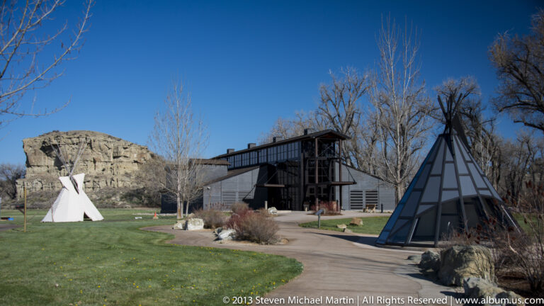 A low visitor center, white conical tent, and glass pyramidal structure sit below a sandstone bluff.