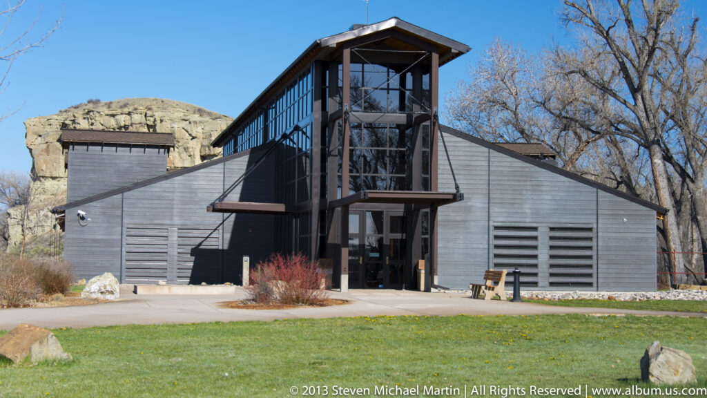 A gray visitor-center building has a tall glass entrance tower and low wings beneath a sandstone bluff.