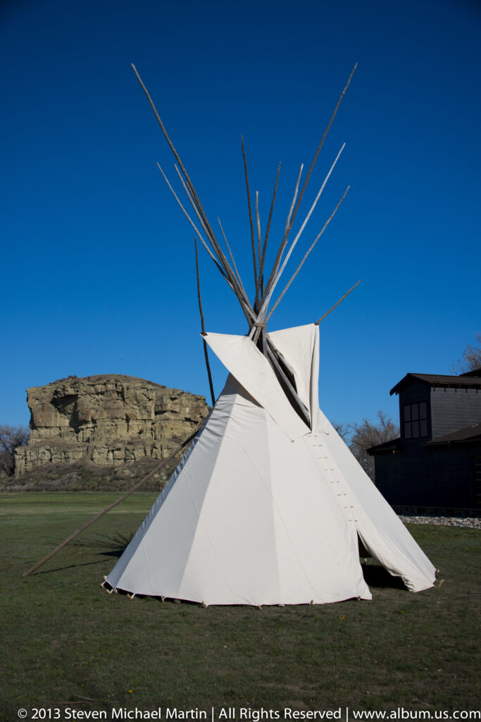 A white conical tent with exposed poles stands on grass in front of a sandstone bluff.