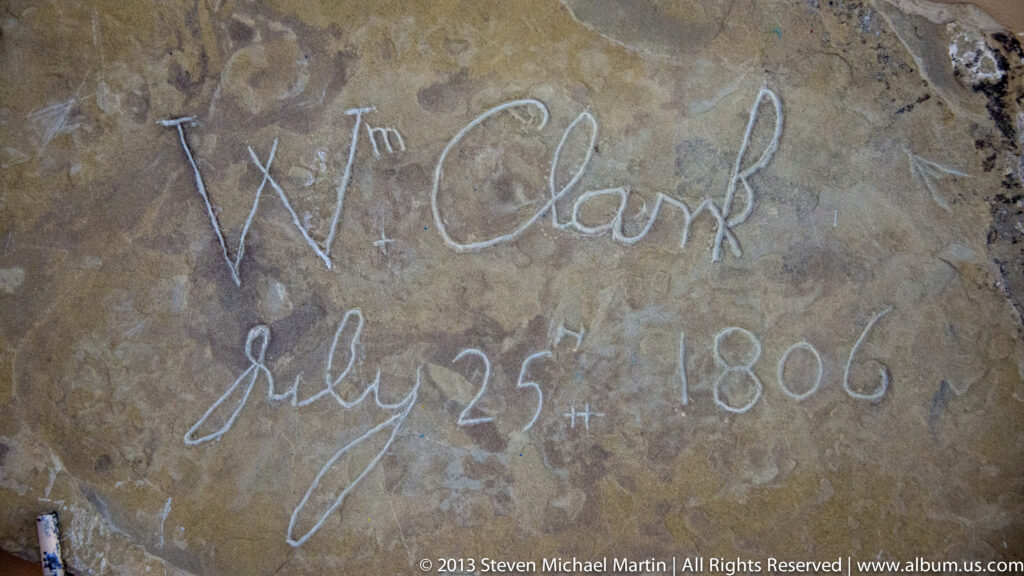 A weathered sandstone face bears a scratched name and date that appear to read “W. Clark” and “July 25th 1806.”