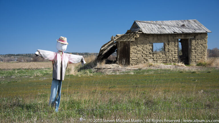 A scarecrow with outstretched arms stands in grass beside a weathered stone building.