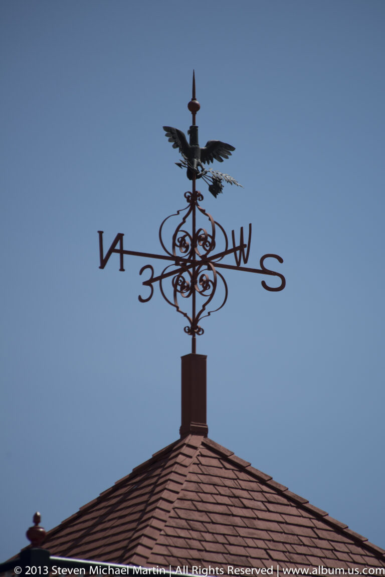An ornate eagle-topped weather vane with compass arms stands above a shingled roof.
