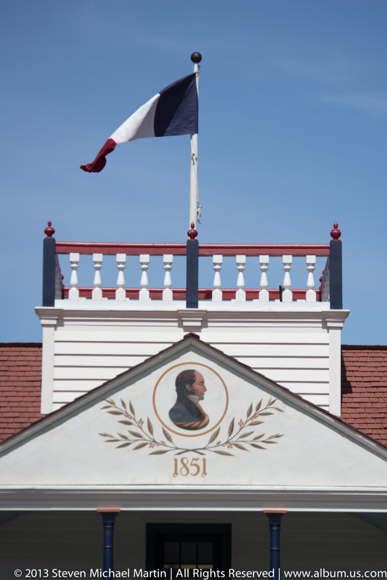 A flag flies above a pediment with a portrait medallion, laurel branches, and the date 1851.