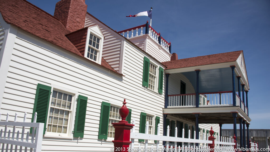A white building with green shutters and an upper gallery stands beside a red pump.