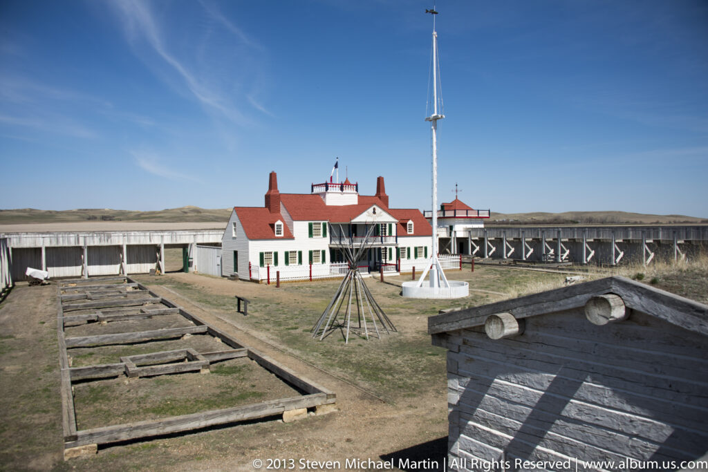 A timber-walled courtyard surrounds white, red-roofed buildings, walkways, and a central flagpole.