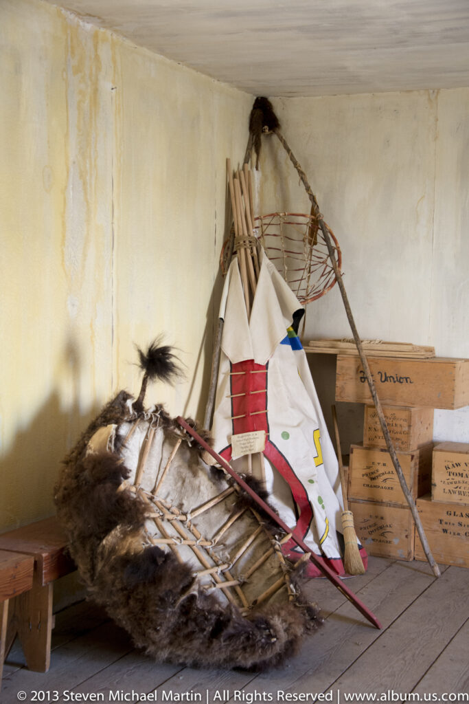 A tipi-style lodge display with poles and hide stands beside a fur-covered structure and crates.