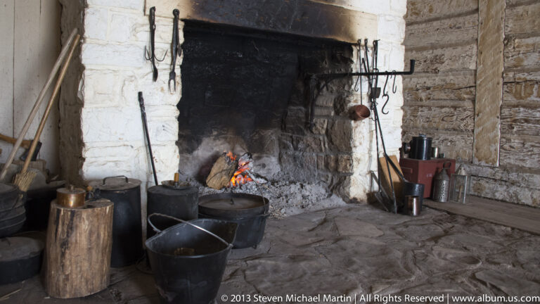 A small fire burns in a stone hearth surrounded by iron pots and hanging fireplace tools.