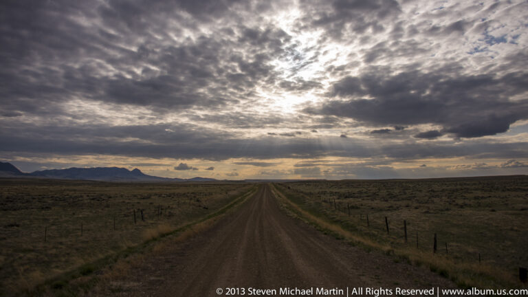 A dirt road crosses open scrubland toward distant hills beneath layered clouds and a bright band of low sunlight.