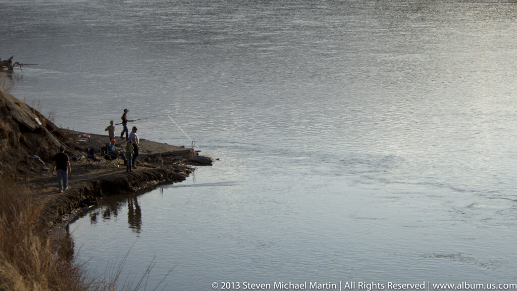 Several anglers gather on an eroded bank, with fishing rods reaching over the broad river.