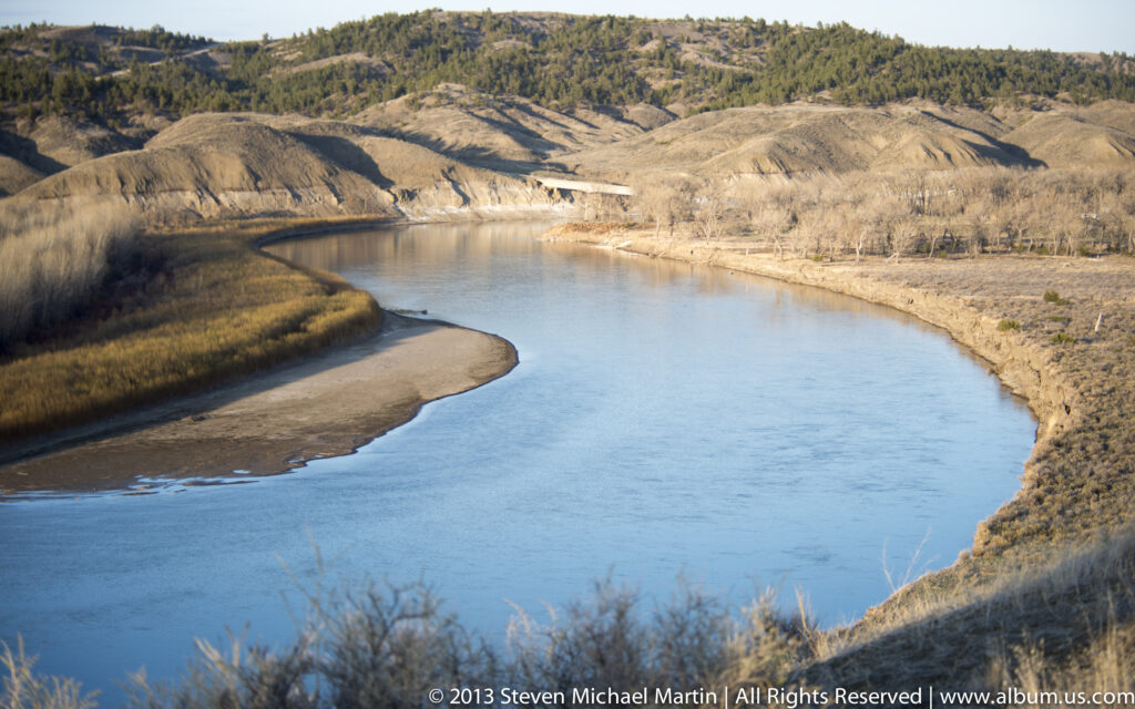 A blue river curves around a pale sandbar below dry hills and leafless riverside trees.