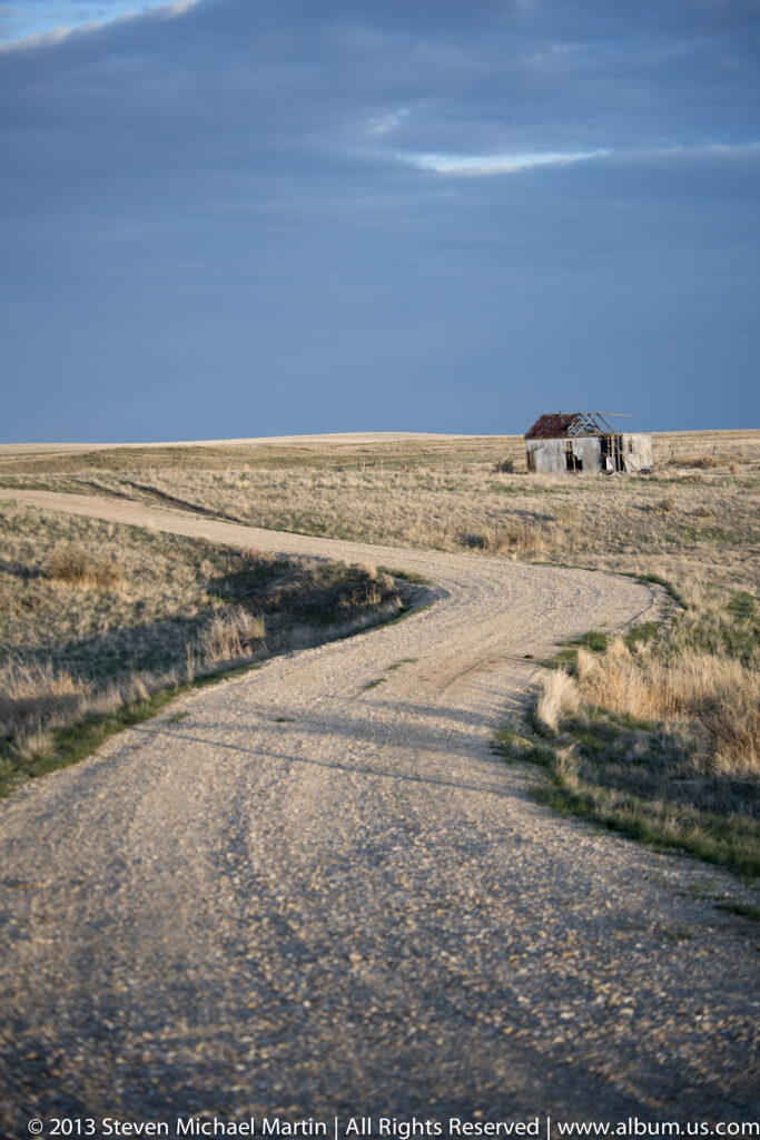 A gravel track crosses dry prairie toward a small, weathered outbuilding.