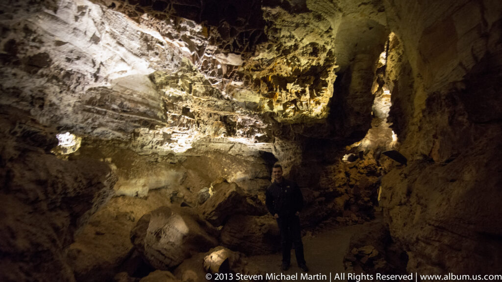 A visitor stands in a broad cave chamber beneath a high, irregularly lit ceiling.