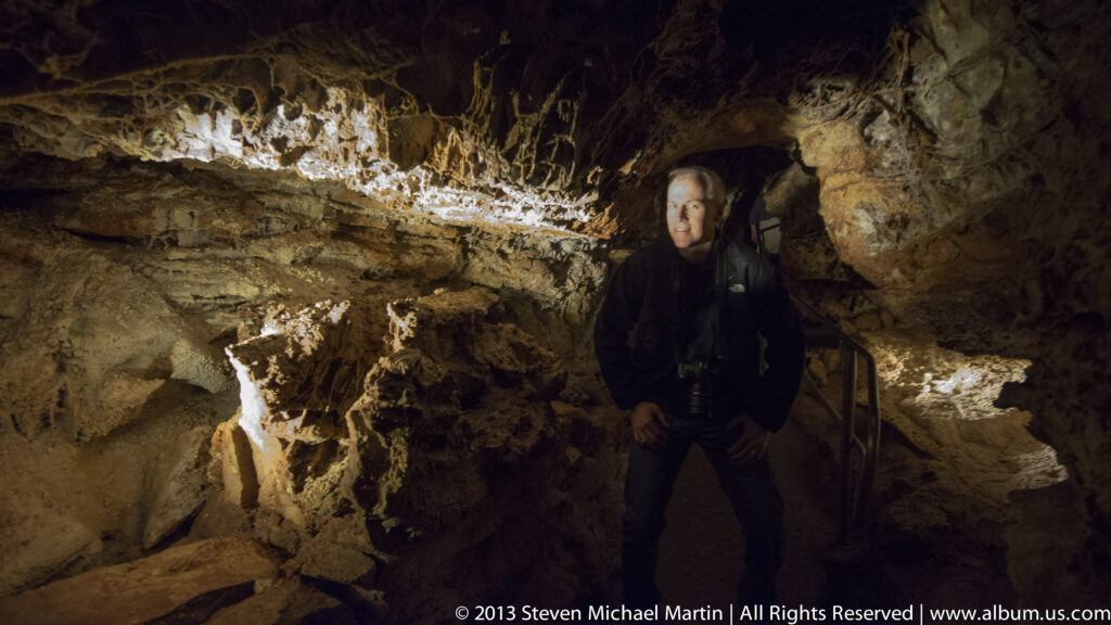 A visitor stands in a shadowed cave passage beneath uneven, warmly lit rock ceilings.