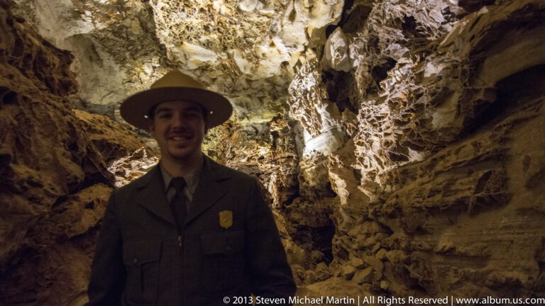 A person in a broad-brimmed hat and dark uniform stands beneath illuminated cave rock formations.