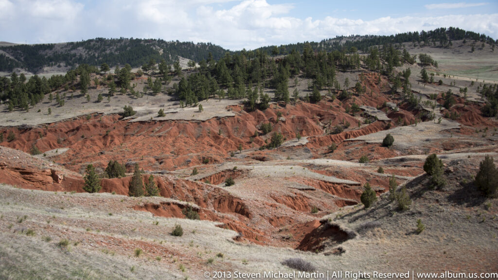 Red and pale ravines cross open slopes below a pine-covered ridge beneath scattered clouds.