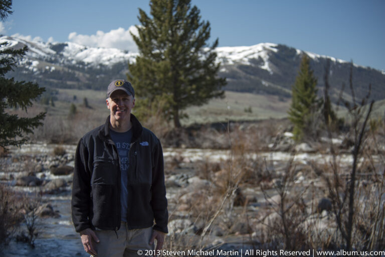 A visitor stands on a snowy path with evergreen trees and pale thermal ground behind him.