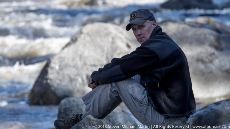 A seated visitor looks back from a rock beside a pale thermal feature and blue water.
