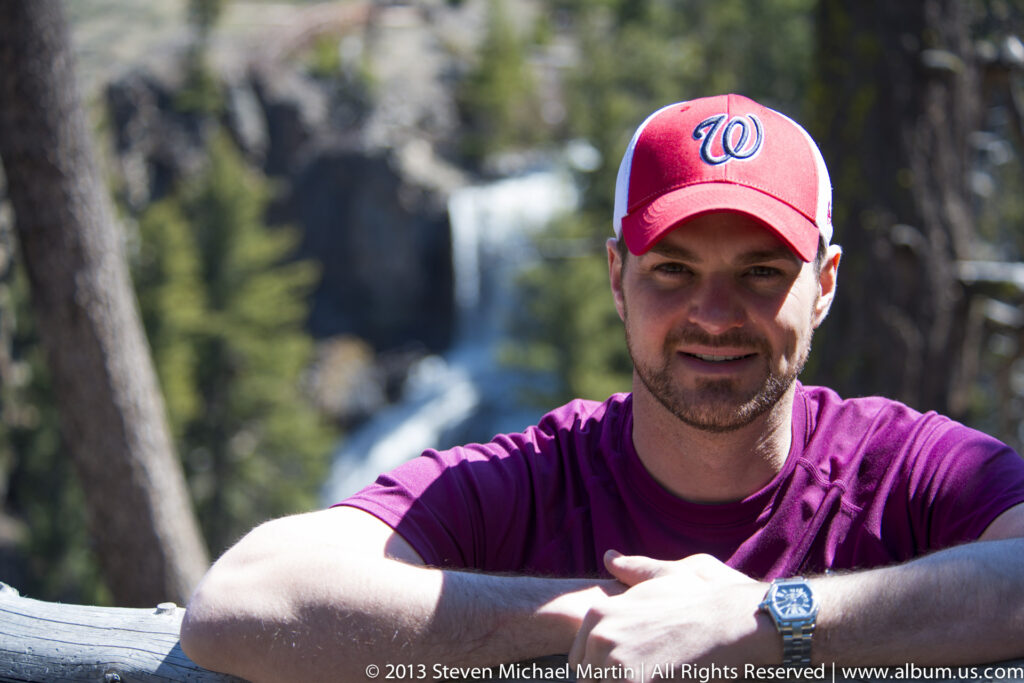 A man in a red cap leans on a wooden rail with a narrow waterfall behind him.
