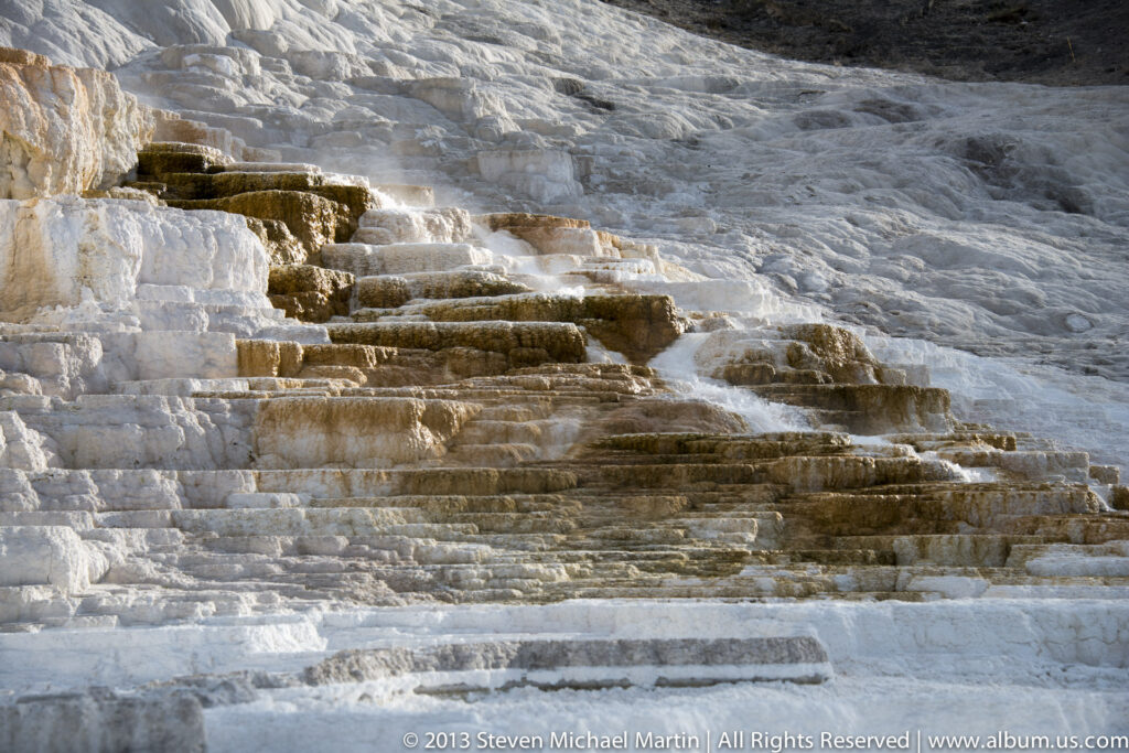 Pale travertine terraces descend in rounded steps across a sunlit geothermal slope.