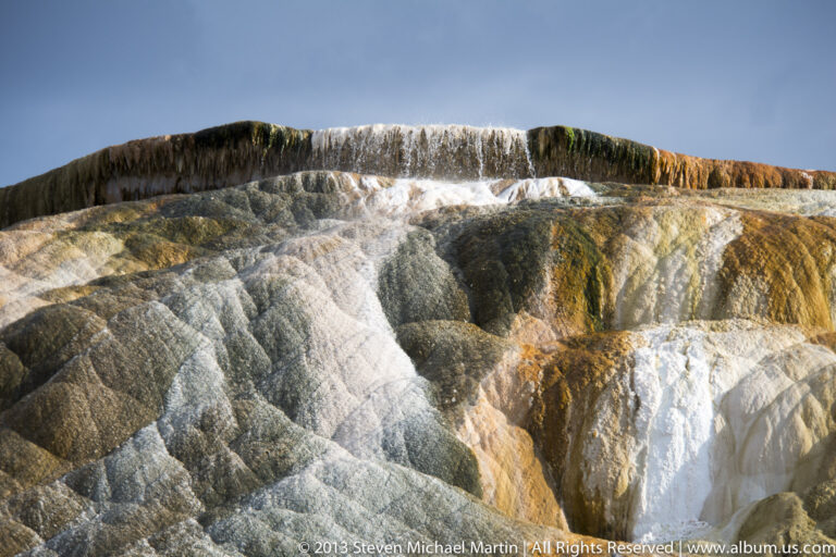 White, gray, ochre, and green bands follow a rounded travertine terrace beneath blue sky.