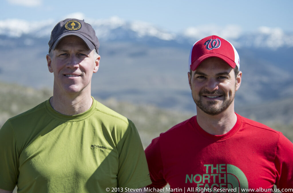 Two men pose together in front of a distant mountain slope beneath a blue sky.