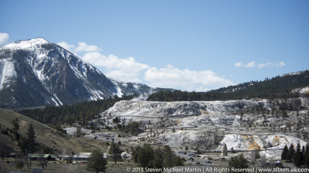Travertine terraces and evergreen forest lie below a snow-streaked mountain ridge in a broad valley.
