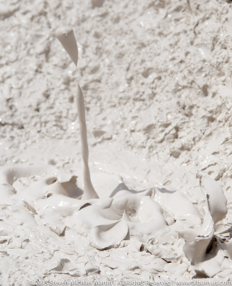 A thin pale plume rises from a small opening in a broad, light-colored geothermal surface.