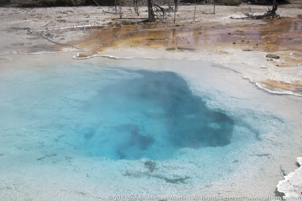A turquoise hot spring fades to pale blue at its mineral rim, with orange mats and sparse trees beyond.