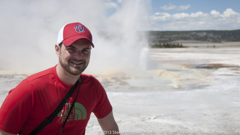 A visitor in a red cap and shirt stands before a steaming geothermal basin.