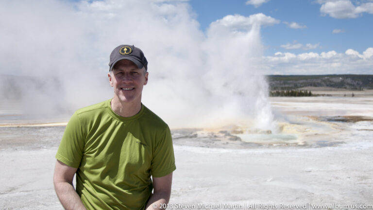 A visitor in a green shirt stands against a large steam plume over a pale geothermal flat.