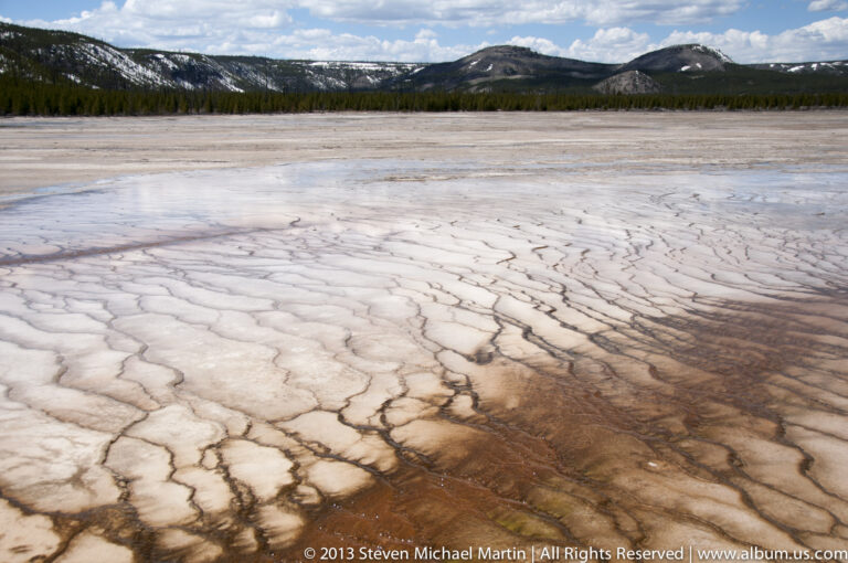 Cracked pale mineral crust forms polygons across a thermal flat, with rust-colored seams and distant hills.