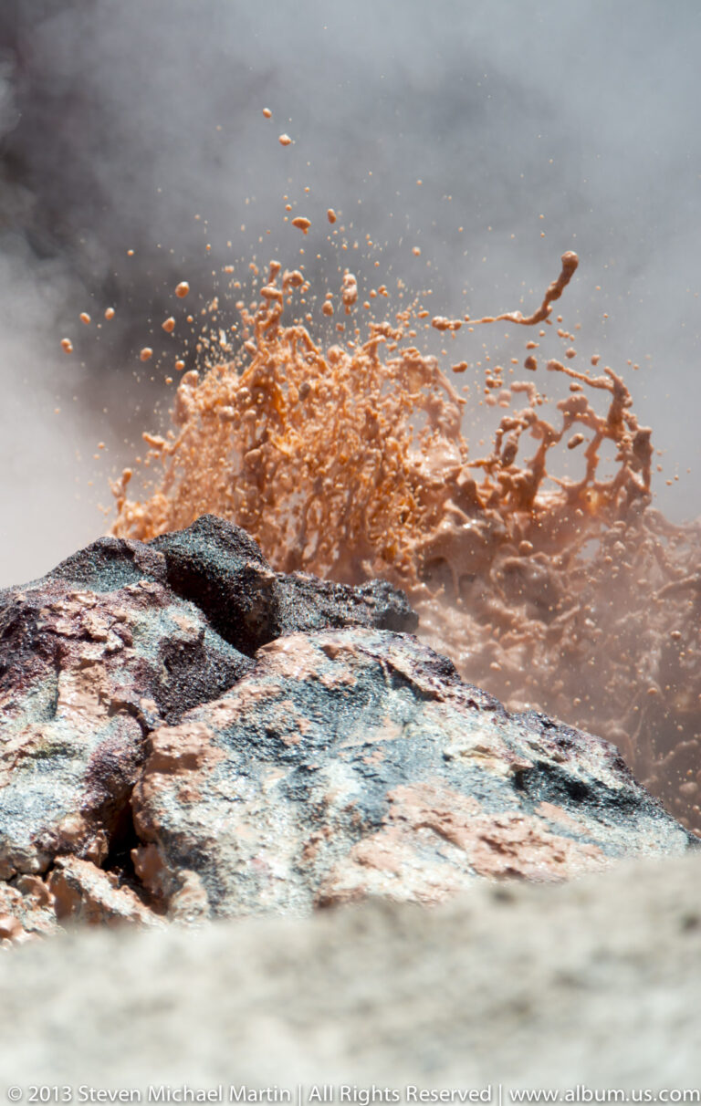 Orange-brown water splashes behind a dark mineral-coated rock ledge in a geothermal pool.