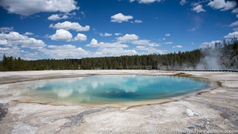 A blue hot spring sits in a pale basin below a boardwalk, trees, and rising steam.