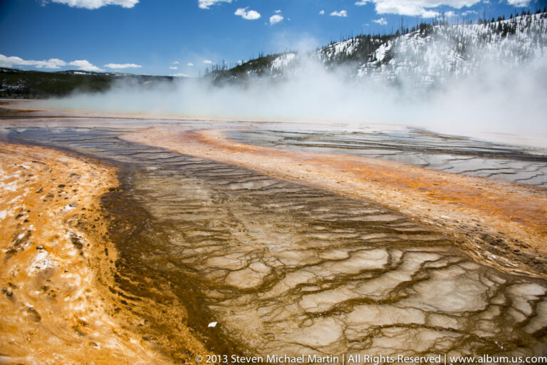 Orange and brown channels cross a pale thermal flat beneath drifting steam.
