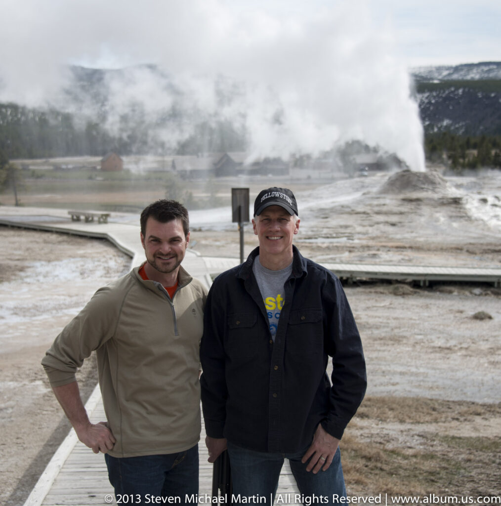 Two men pose beside a boardwalk with steaming geothermal features behind them.