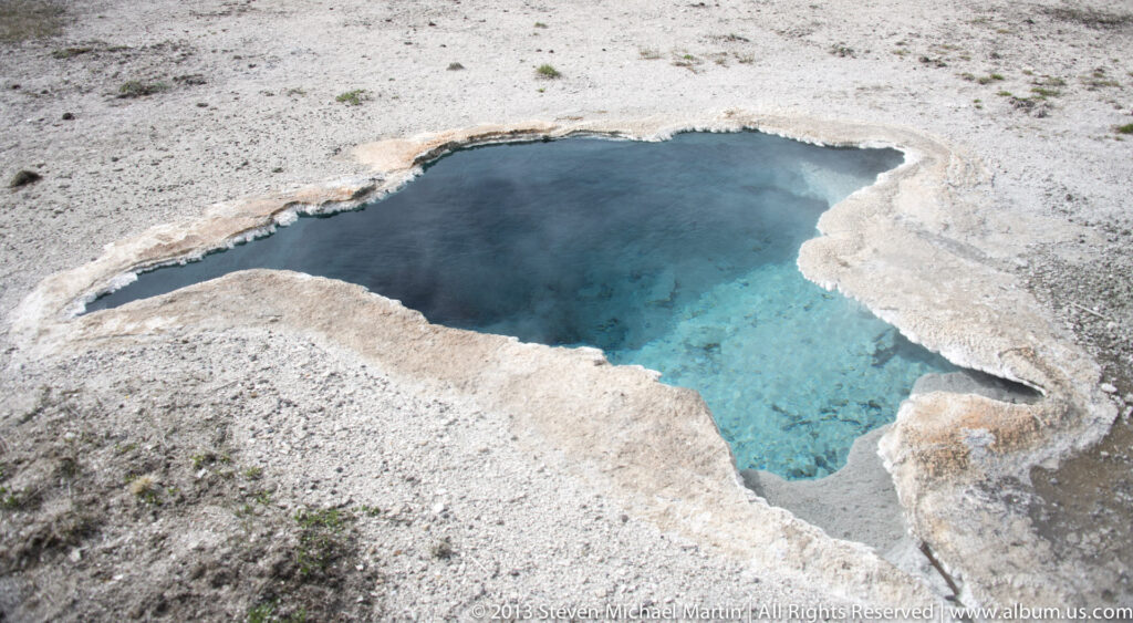 A small turquoise pool is ringed by rough white mineral crust on a pale geothermal surface.